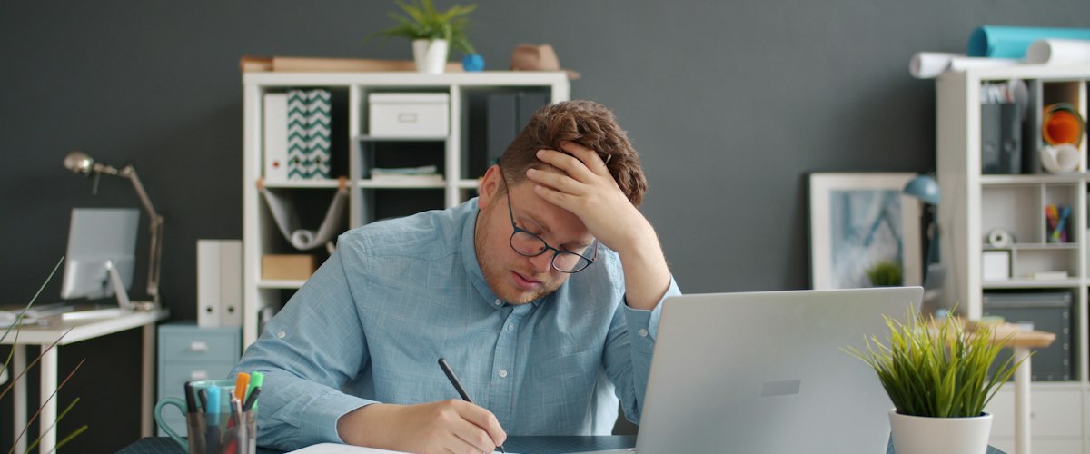 Man writing at desk with laptop, looking stressed