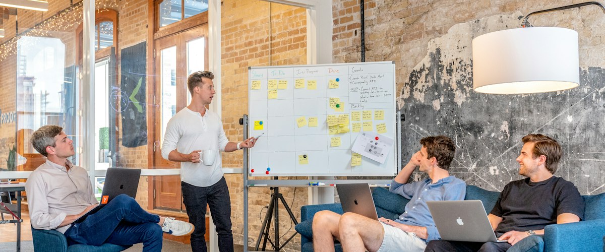 Three men sitting while using laptops and watching man beside whiteboard